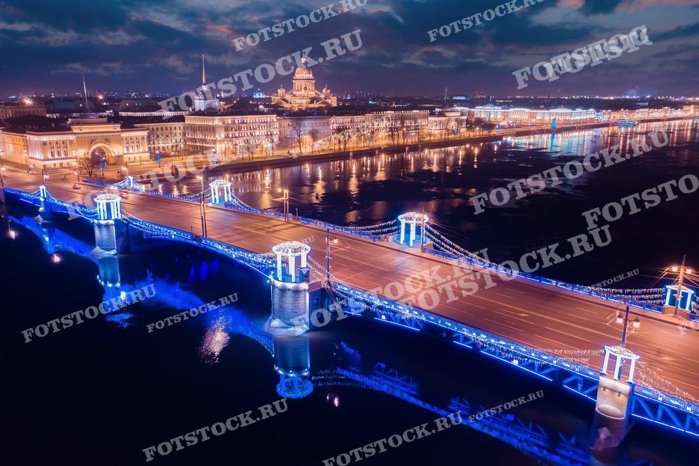 На фотографии изображён ночной вид Санкт-Петербурга с высоты птичьего полёта. Ярко освещённый Дворцовый мост перекинут через реку Неву, соединяя две части города. На заднем плане видны здания, подсвеченные разноцветными огнями, отражающие архитектурную красоту и историческое наследие города. 
Это фото может быть использовано для иллюстрации туристических статей о Санкт-Петербурге, в печатных и электронных путеводителях, а также в качестве рекламного материала для агентств, организующих поездки в этот город. Фотография также подойдёт для украшения интерьеров или создания тематических коллажей.